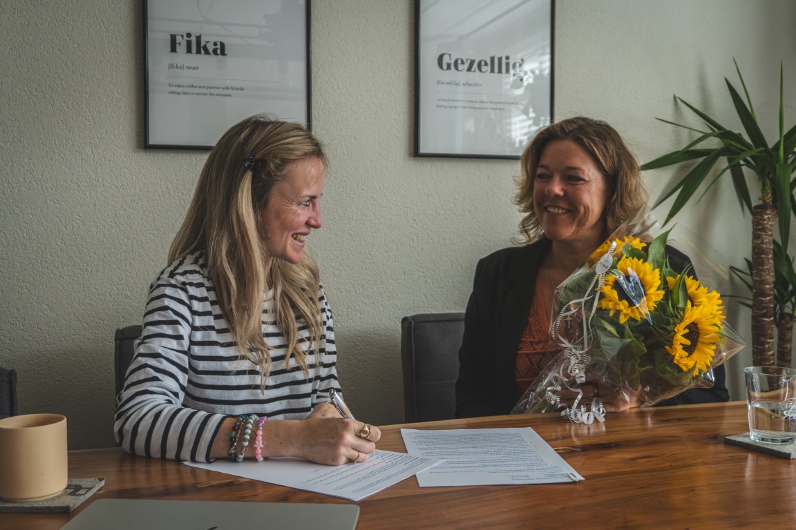 Celeste tekent een boekcontract en uitgever Jet Hoogerwaard zit ernaast met een bos bloemen