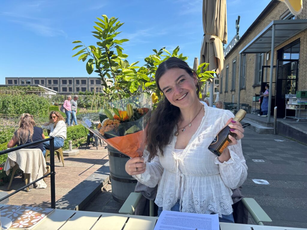 nne Bootz zit buiten op een terras en draagt een witte blouse. Ze houdt een fles champagne in haar rechterhand en een boeket bloemen in haar linkerhand. Voor haar op de tafel ligt een document. Op de achtergrond zijn mensen te zien die aan tafels zitten, met een gebouw en een terras. Het is een zonnige dag met een blauwe lucht.